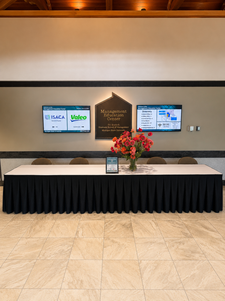 A fron view of the entrance table with flowers in a vase, the placard, and two TV screens.